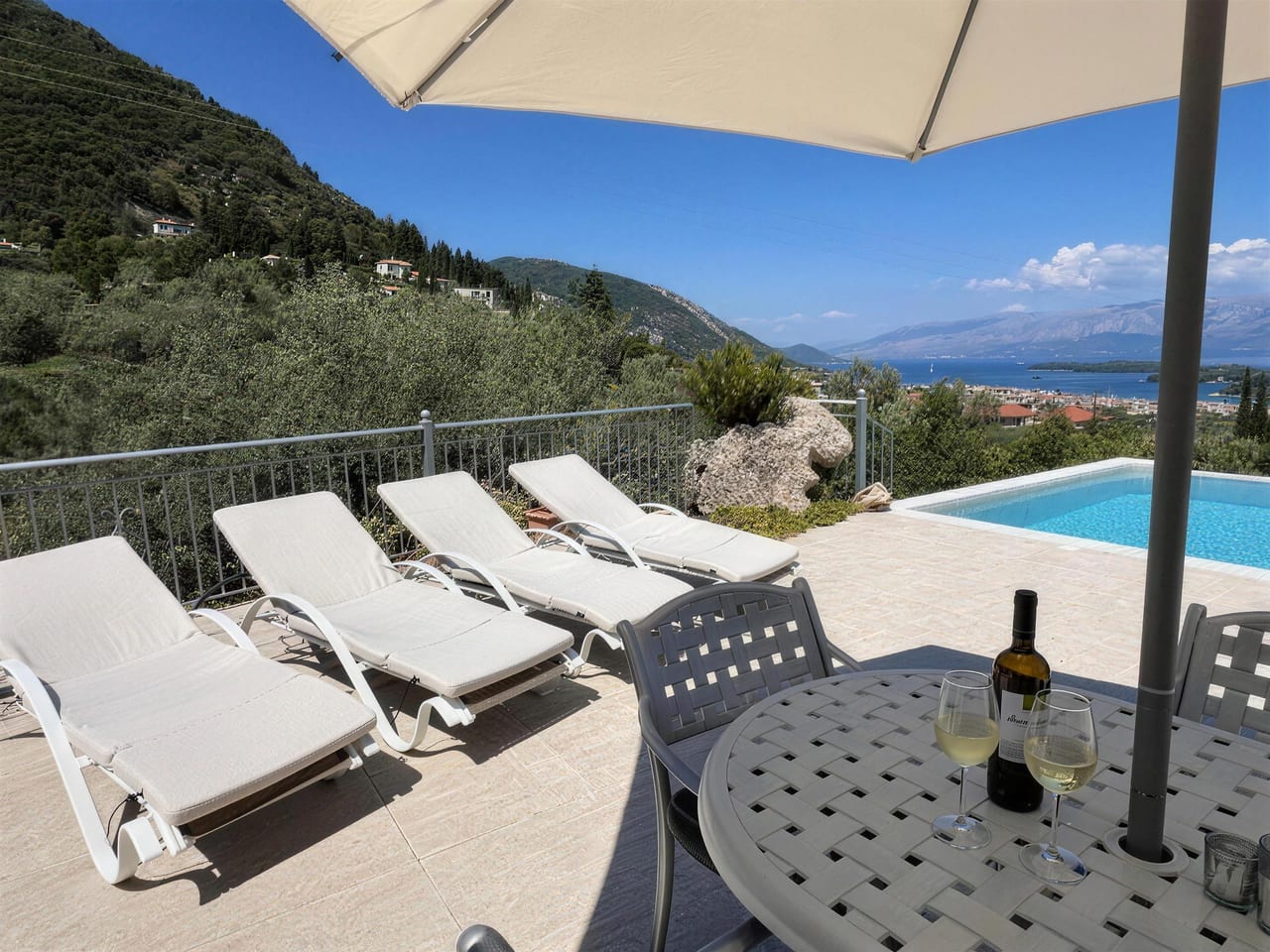 Loungers and a shaded table on the pool terrace, the bay and mountains beyond